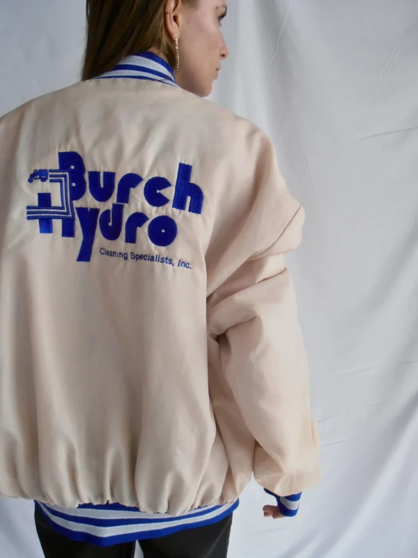 Back view of a vintage beige bomber jacket with blue and white striped cuffs, collar, and waistband, and blue lettering on the back.