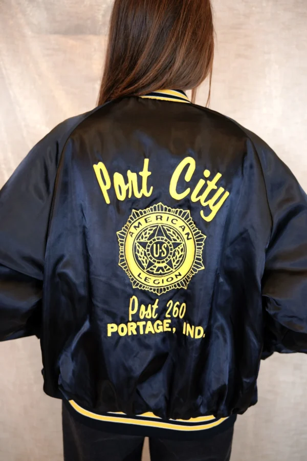 Back view of a vintage black bomber jacket with yellow lettering and design on the back.
